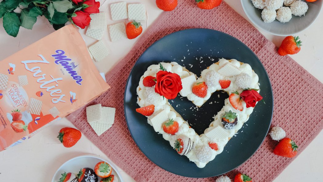 Muttertagstorte im Letter Cake Stil auf grauem Teller. Daneben befinden sich Erdbeeren, Manner Waffeln und rote Rosen