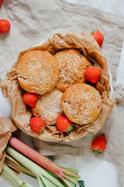 Saftige Erdbeer-Rhabarber-Muffins mit Kokos. Daneben liegen Erdbeeren und frischer Rhabarber verteilt.